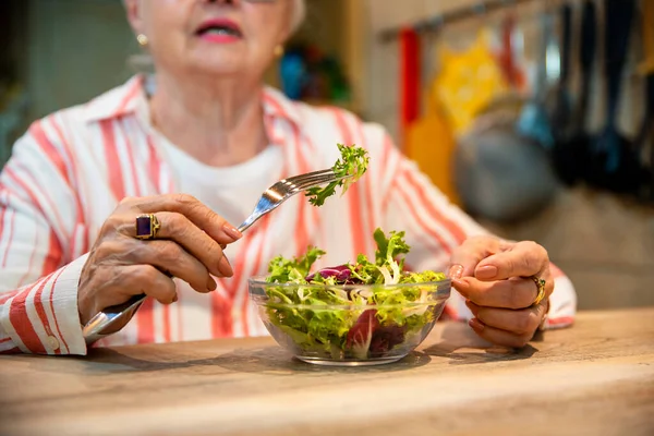 Sağlıklı atıştırmalıklar ve bir bardak su içen yaşlı kadın. Yaşlı kadın ahşap mutfak masasında taze salata yiyor. Boşluğu kopyala