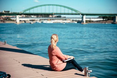 İnternette çalışan dijital kadın göçebe nehir rıhtımında oturuyor. Şehir nehir kıyısında dizüstü bilgisayar kullanan sarışın bir kadın. Boşluğu kopyala