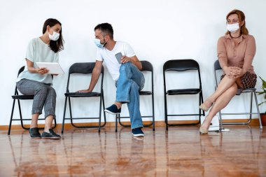 İnsanlar sıraya dizilmiş sandalyelere oturuyor. Bekleme odasında çok ırklı insanlar var.