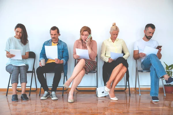 İnsanlar sıraya dizilmiş sandalyelere oturuyor. Bekleme odasında çok ırklı insanlar var.