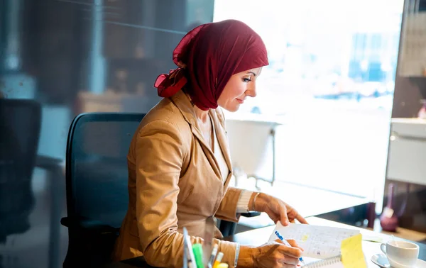 Arabic Woman secretary at work, sitting at office desk. Portrait of Islamic female assistant wearing hijab at workplace. Arab Woman with red abaya scarf in Company job