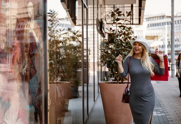 Woman shopping in the city, looking at store window. Pretty blonde long hair female with bags shopping in season sale