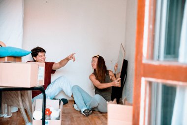 Yeni dairenin zemininde ev eşyalarıyla oturan iki genç insan. Çift taşınıyor. Yeni evde paketler açılıyor