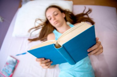 Yatakta uzanırken elinde kitapla gülümseyen genç tatlı kız. Okul ödevi için kitap okuyorum. Görünümün üstünde