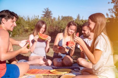 Gençler yaz günü dışarıda piknik yaparken karpuz dilimleriyle tezahürat yapıyorlar. Gençler doğada birlikte eğleniyor, taze meyve yiyorlar.