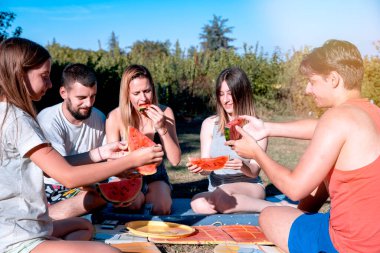 Gençler yaz günü dışarıda piknik yaparken karpuz dilimleriyle tezahürat yapıyorlar. Gençler doğada birlikte eğleniyor, taze meyve yiyorlar.