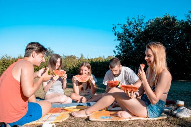 Gençler yaz günü dışarıda piknik yaparken karpuz dilimleriyle tezahürat yapıyorlar. Gençler doğada birlikte eğleniyor, taze meyve yiyorlar.