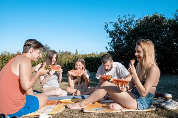 Young People cheering with Watermelon slices having Picnic outdoors on Summer day. Teenagers having fun together in Nature, eating fresh fruit