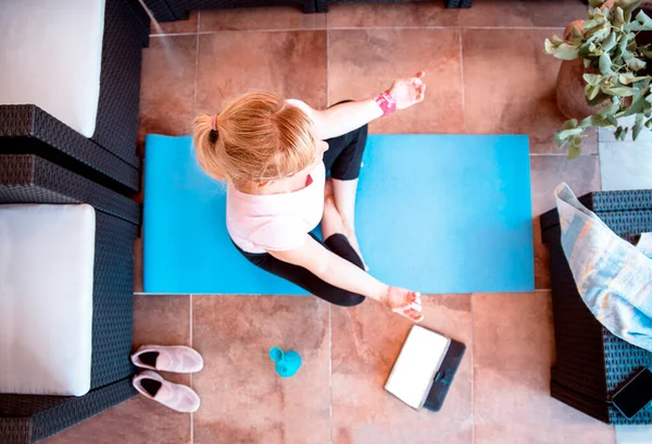 Woman meditate watching video tutorial online on tablet. Mid adult Female doing Yoga workout at home sitting on mat in living room