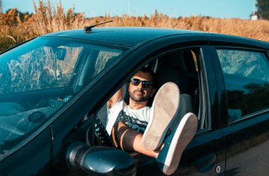 Güneşli bir günde araba şoförü koltuğunda dinlenen güneş gözlüklü yakışıklı sakallı adam. Tatilde bir yol gezisinde bacakların pencerenin önünde dinleniyor.