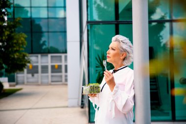 Yemek molasında güzel, gri saçlı bir kadın. Taze salata yiyen güzel yaşlı kadın. Ofis binasının önünde salata yiyen yaşlı kadın. Boşluğu kopyala