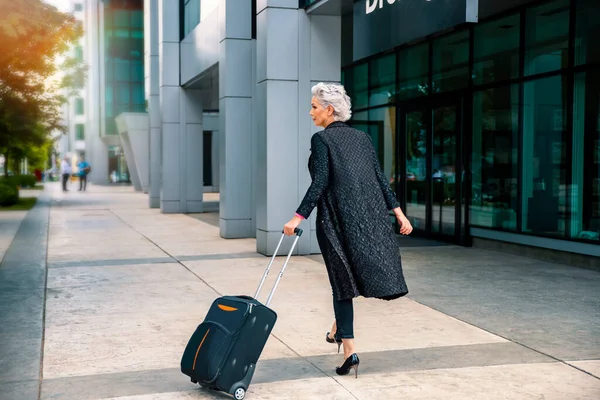 Elderly white hair female on heels walking with luggage to airport. Woman traveler in black going to business trip, copy space