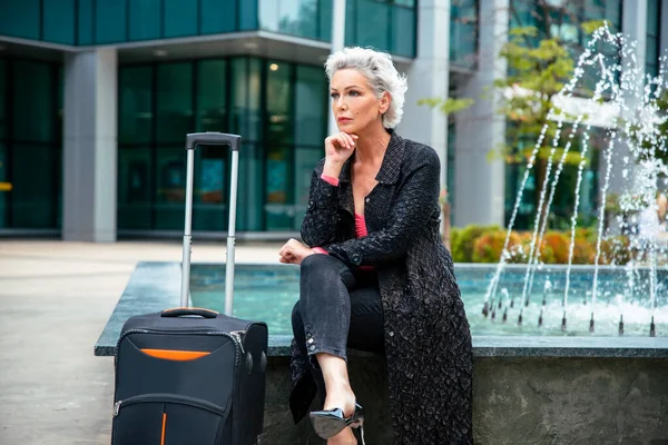 Elderly white hair female on heels walking with luggage to airport. Woman traveler in black going to business trip, copy space