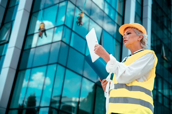 Daha yaşlı, gri saçlı bir mimar. İnşaat alanının planlarını kontrol ediyor. Sarı başlıklı ve yansıtıcı yeleği olan yaşlı kadın inşaat mühendisi. Boşluğu kopyala