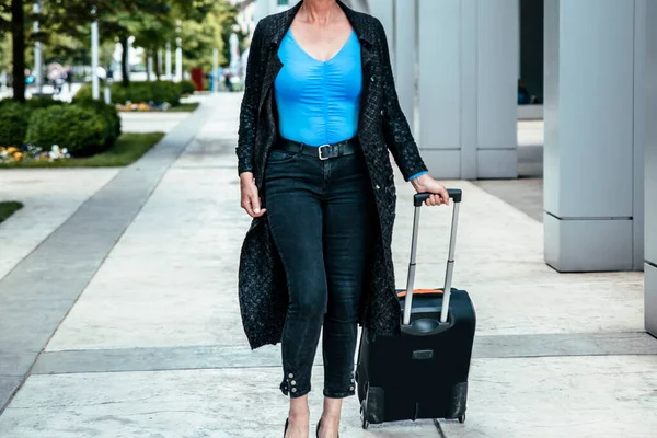 Elderly white hair female on heels walking with luggage to airport. Woman traveler in black going to business trip, copy space