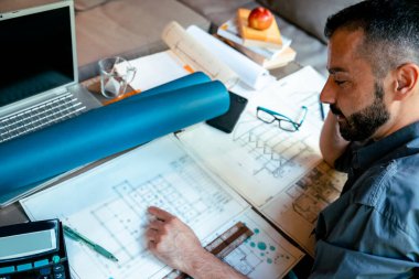 Erkek mimar yeni emlak projesini çiziyor. Sakallı, gülümseyen erkek inşaat mühendisi masasında planlar üzerinde çalışıyor. Seçici odak