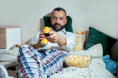 Changing the TV channel using remote control. Man laying in bed drinking beer can and eating popcorns. Guy in pajamas enjoying the sports game at home