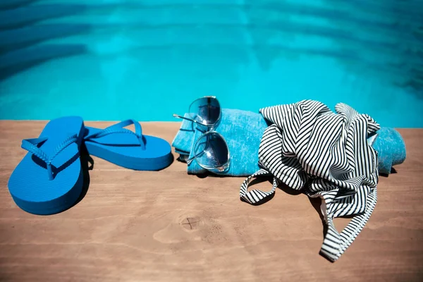 Summer Vacation accessories on the pool beach. Blue Flip flops, sunglasses, swimsuit and towel on the wooden poolside. Spa Holiday concept. Copy space