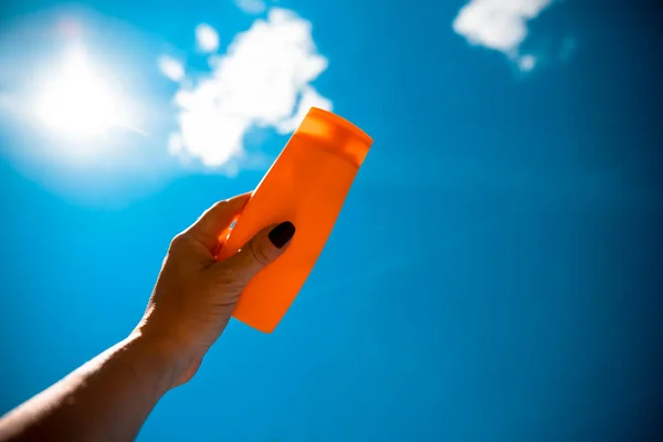 Sunscreen lotion orange bottle in female hands against blue sunny sky. Sun tanning with Sunblock protection. Skin care on summer day background. Copy space for text