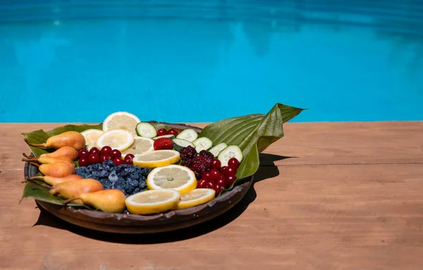 Havuz kenarındaki ahşap tabakta renkli meyve karışımı. Lüks tatilde sağlıklı atıştırmalıklar. Yaz tatilinde taze meyve yemek yüzme havuzuna mavi su satın almak. Boşluğu Kopyala