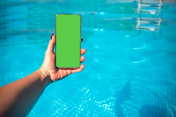 Smartphone with chroma key green screen in female hand against blue pool water. Advertising background