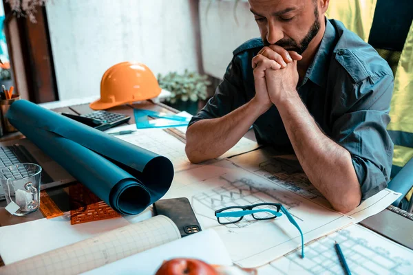 Overworked stressed Man engineer having headache while working on his project blueprint at work. Frustrated male with tension sitting at desk full of drawings