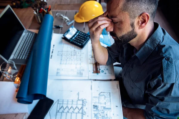 Overworked stressed Man engineer having headache while working on his project blueprint at work. Frustrated male with tension sitting at desk full of drawings