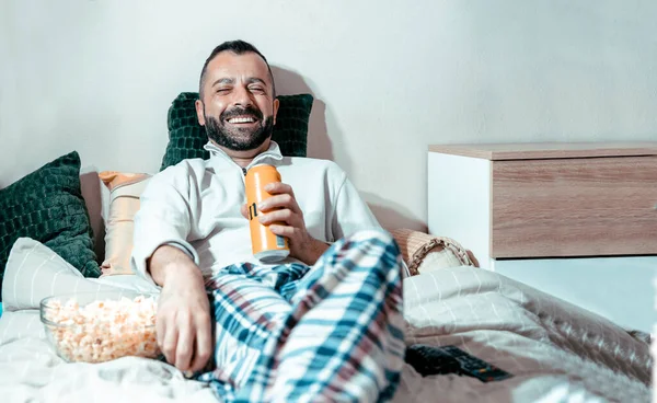 Happy Man watching television while laying in bed with yellow beer can and popcorns. Enjoying in online TV show. Handsome Guy in pajamas looking sports game at home