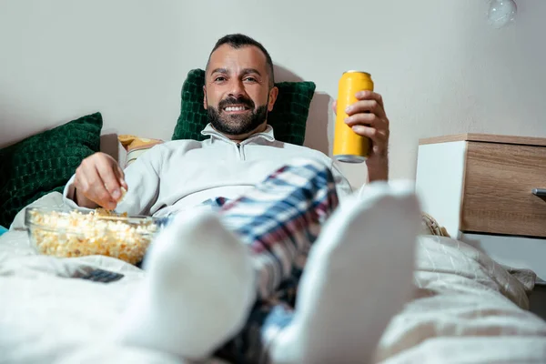 Happy Man Ogląda Telewizję Leżąc Łóżku Żółtą Puszką Piwa Popcornami — Zdjęcie stockowe