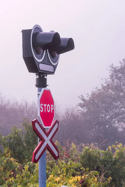 Railroad crossing with warning signs and lights and stop sign below. In foggy day with autumn field