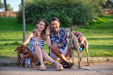 Genç, dövmeli bir çift köpekleriyle oturup kucaklaşıyor ve kameraya bakıyor..