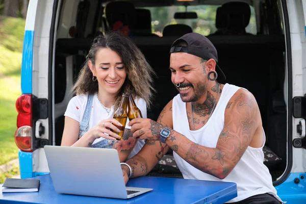 Jovem casal tatuado sorrir, fazer um brinde com cerveja em uma chamada de vídeo laptop. — Fotografia de Stock