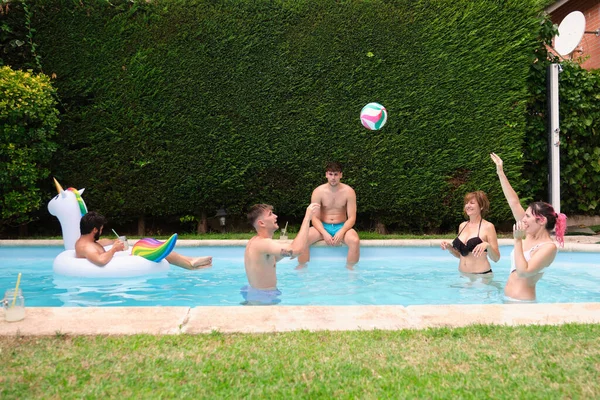 Bir grup insan bir top ve gökkuşağı tek boynuzlu atıyla havuzda yüzüyor..