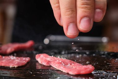 Sıcak taşın üstünde ızgara bifteğe tuz ekleyen bir eli kapat..