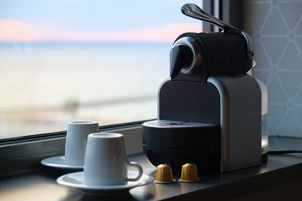 Cafetera con cápsulas y dos tazas listas para usar cerca de una ventana. — Foto de Stock