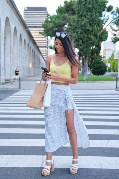 Mladý latina žena pomocí telefonu chůze držení nákupních tašek. — Stock fotografie