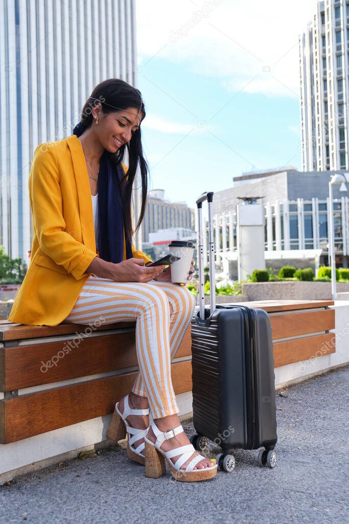 Young indian businesswoman using a phone, with a cup of coffee and a suitcase.