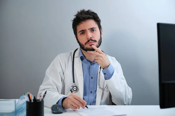 Young doctor looking at camera and thinking sitting at desk in a medical office. — Fotografia de Stock