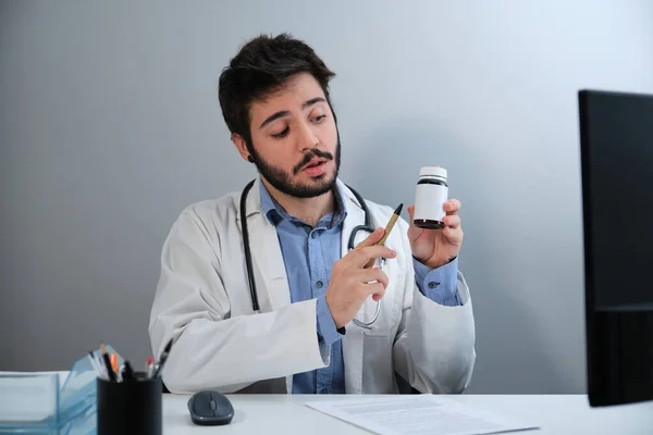 Ung läkare prata och peka på en piller flaska sitter vid skrivbordet på läkarmottagningen. — Stockfoto
