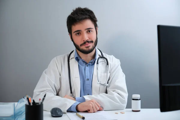 젊은 의사가 의료 실에서 카메라를 보며 책상에 앉아서 웃고 있는 모습. — 스톡 사진