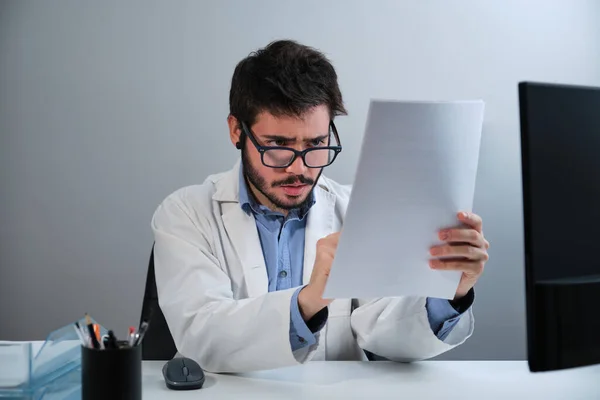 Ung läkare tittar på den medicinska rapporten sitter vid skrivbordet på ett läkarkontor. — Stockfoto