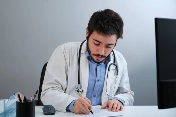 Ung läkare skriver en medicinsk rapport sitter vid skrivbordet på ett läkarkontor. — Stockfoto