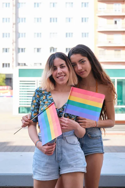Giovane coppia lesbica abbracciare, tenendo bandiere arcobaleno e guardando la fotocamera. — Foto Stock