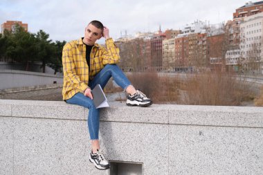 Duvarda oturmuş, elinde bir kitapla kameraya bakan iki kişilikli olmayan bir genç..