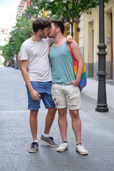 Gay pár smích a objímání sedí na lavičce drží LGBT vlajky. — Stock fotografie