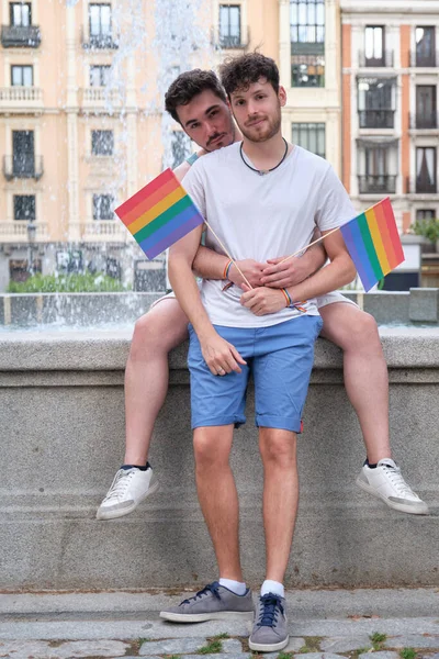 Couple gay étreignant et regardant la caméra tenant des drapeaux LGBT assis par une police. — Photo