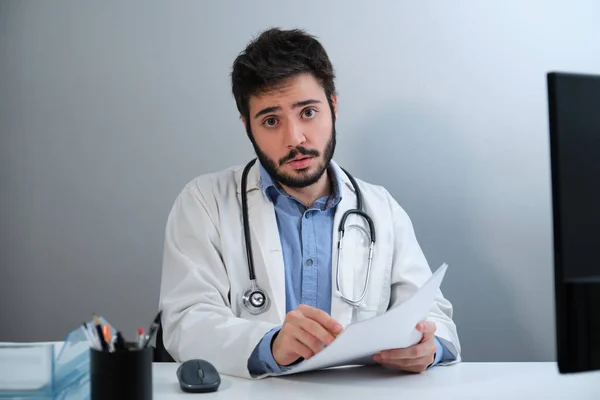 Jovem médico olhando para a câmera e falando sentado na mesa em um consultório médico. — Fotografia de Stock