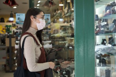 Yüzü maskeli genç Asyalı bir kadın ayakkabı mağazasının vitrinine bakıyor..
