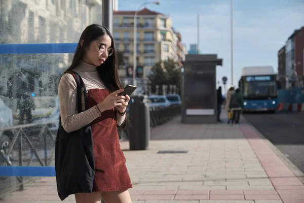 Молода азіатська жінка використовує свій смартфон під час очікування автобусної зупинки . — стокове фото