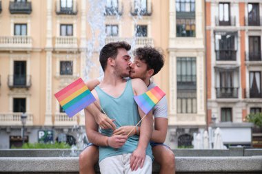 Eşcinsel bir çift bankta oturmuş gülüyor ve sarılıyor ellerinde LGBT bayrakları.